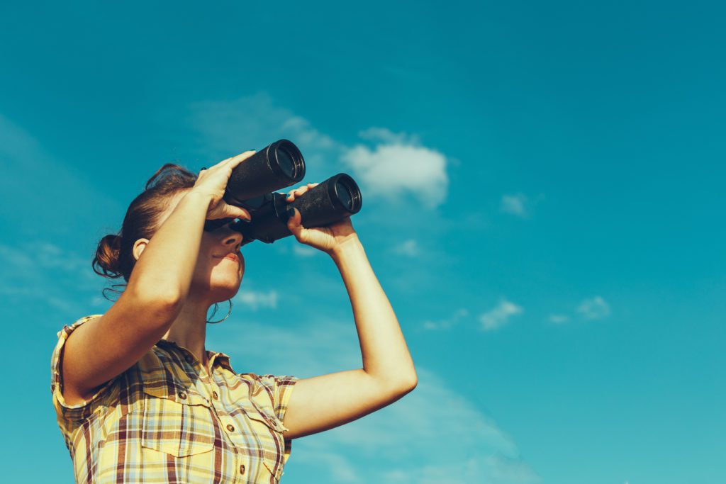 Beautiful young girl looks through binoculars on a bright sunny day, low angle